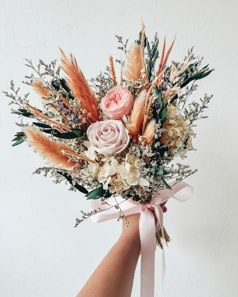 Bouquet of flowers with pink and white roses, pampas grass, and other greenery held by a hand against a light background.
