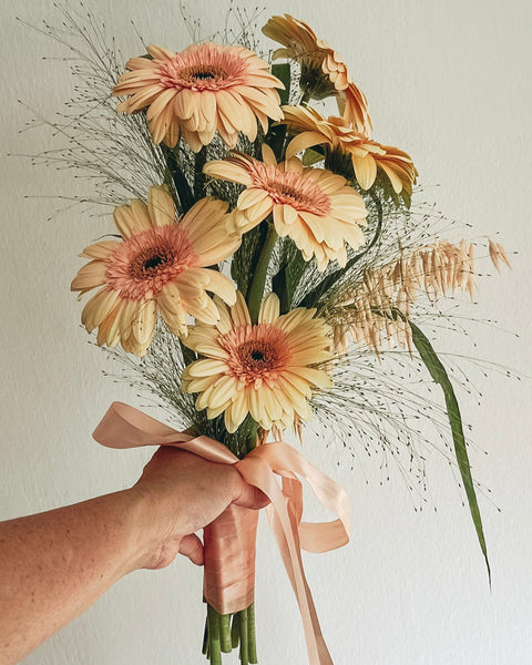 Bouquet of yellow and peach-colored flowers with a pink ribbon held by a hand against a neutral background